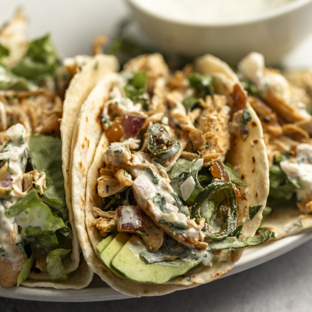 A closeup shot of a tortilla stuffed with shredded chicken, lettuce, avocado slices, all drizzled with a creamy sauce. Three tacos rest on a plate with a small bowl in the background.