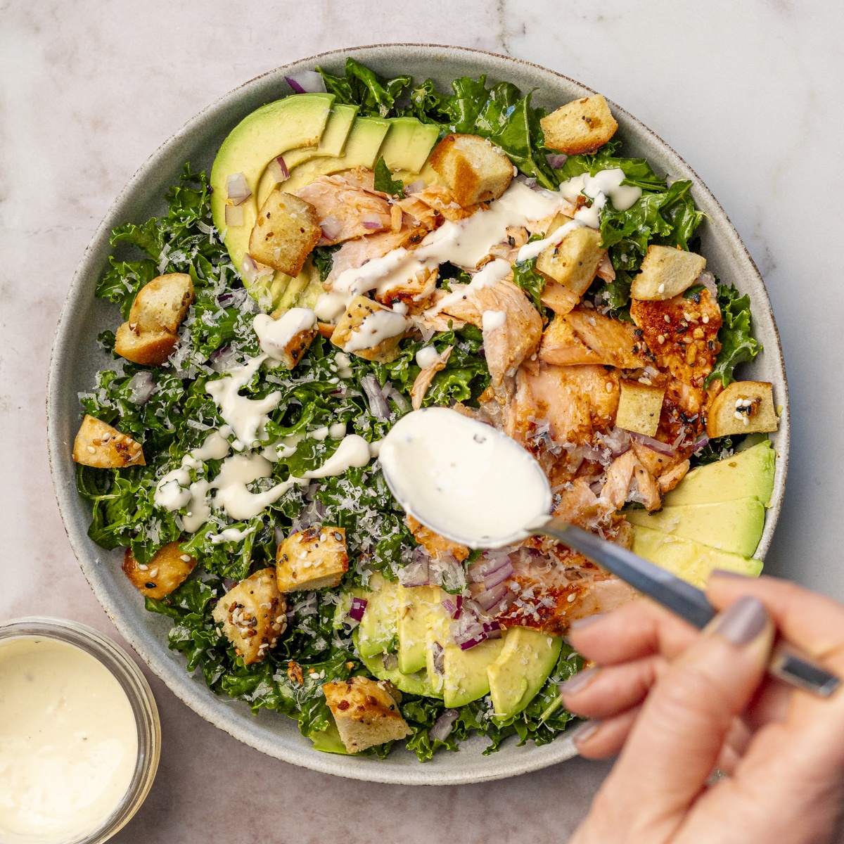 A woman's hand holding a spoon and drizzling dressing over a ceramic bowl full of salmon kale salad. A small bowl of dressing is sitting nearby.